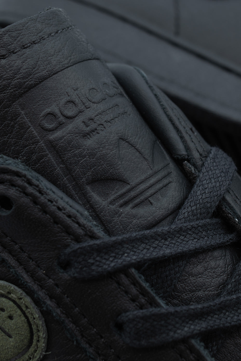 Macro studio close-up of the smooth black leather tongue on the Adidas Superstar ADV Mark Gonzales, displaying the cleanly debossed Adidas Originals Trefoil logo emblem and textured waxed black shoelaces.