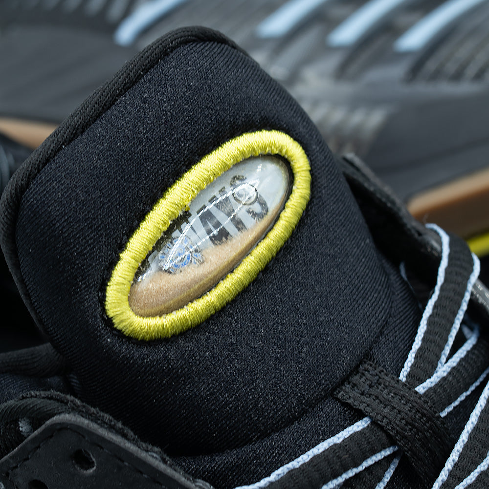 Detailed macro studio close-up of the tongue badge on the adidas x ADWYSD Climacool 1, focusing on the clear oval bubble capsule filled with genuine beach sand and liquid displaying floating ADWYSD text, enclosed within dense neon yellow satin-stitch embroidery over black padded neoprene.