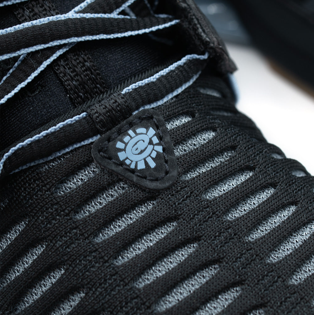 Extreme macro close-up of the lower lacing zone on the adidas x ADWYSD Climacool 1, showcasing the triangular black nubuck lace anchor embroidered with a blue ADWYSD @Sun icon, two-tone rope laces, and the dense vertical knit pattern of the open Climacool breathable mesh.