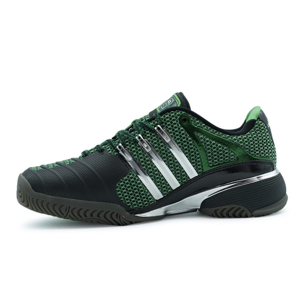 Medial side studio profile of the adidas x Brain Dead Barricade V, showing the extensive matte carbon-black textured rubber toe-drag protection guard, breathable green sandwich mesh midfoot underlay, polished silver three stripes, and chrome midfoot stabilization plate.