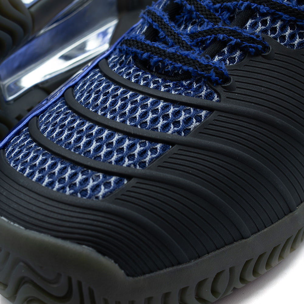 Detailed macro perspective of the forefoot on the adidas x Brain Dead Barricade V, focusing on the Mystery Ink blue open-structure mesh overlaid with grooved black rubber flex guards and the rugged wrapped toe bumper.