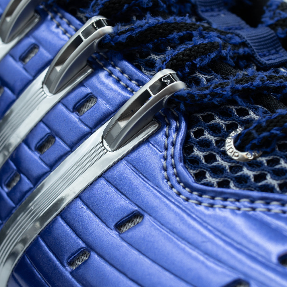 Close-up studio perspective of the lacing system on the adidas x Brain Dead Barricade V, showing the polished chrome Three Stripes cage extending into eyelet anchors, metallic eyelets stamped with "adidas", and two-tone textured blue and black woven laces over mesh.