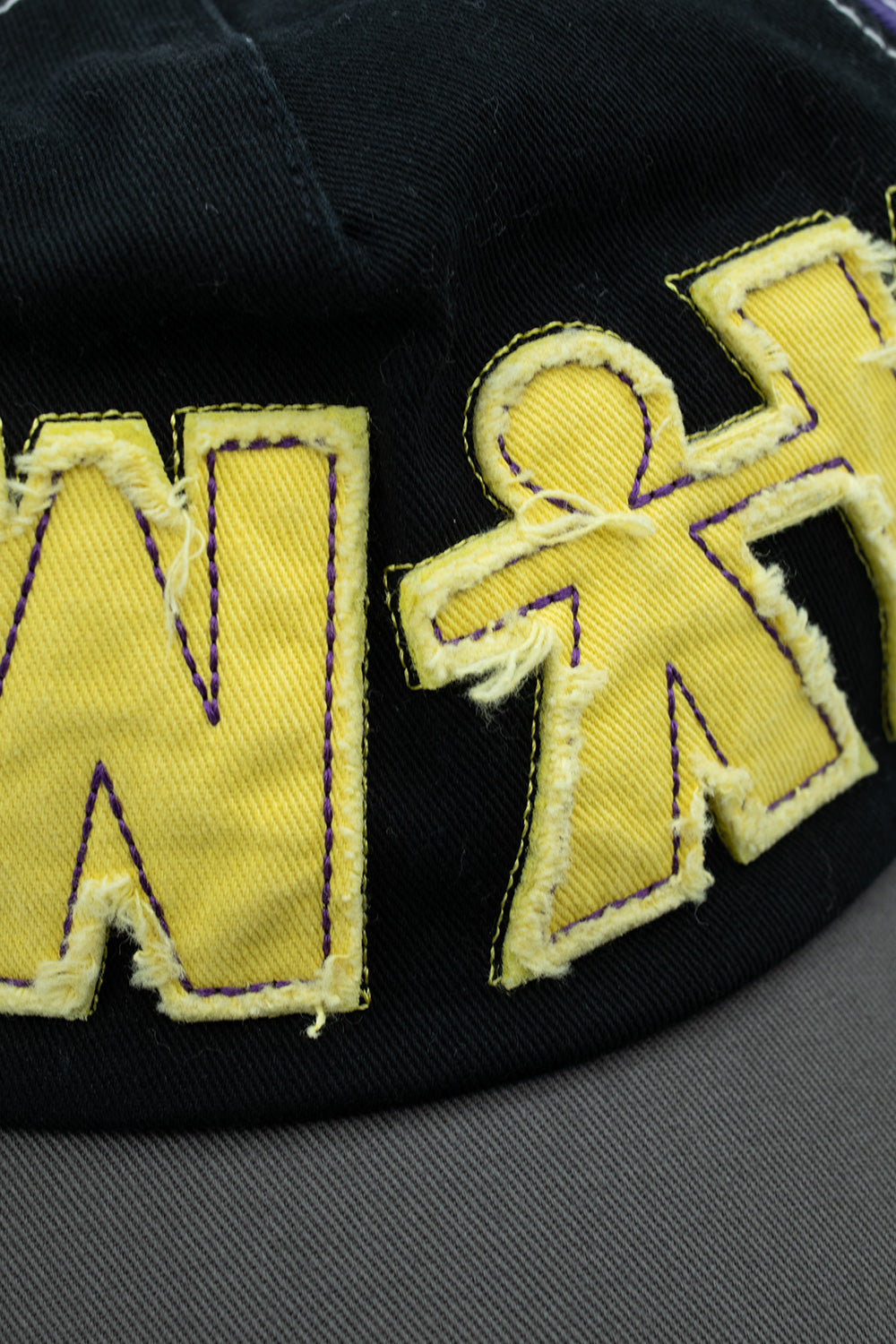 Macro close-up detail shot of the yellow frayed appliqué patches on the black cotton front panel of the Always 5-panel cap, showcasing the textured raw-edge cotton twill, black perimeter border stitching, purple interior contrast thread lines, and the stylized stickman human shape.