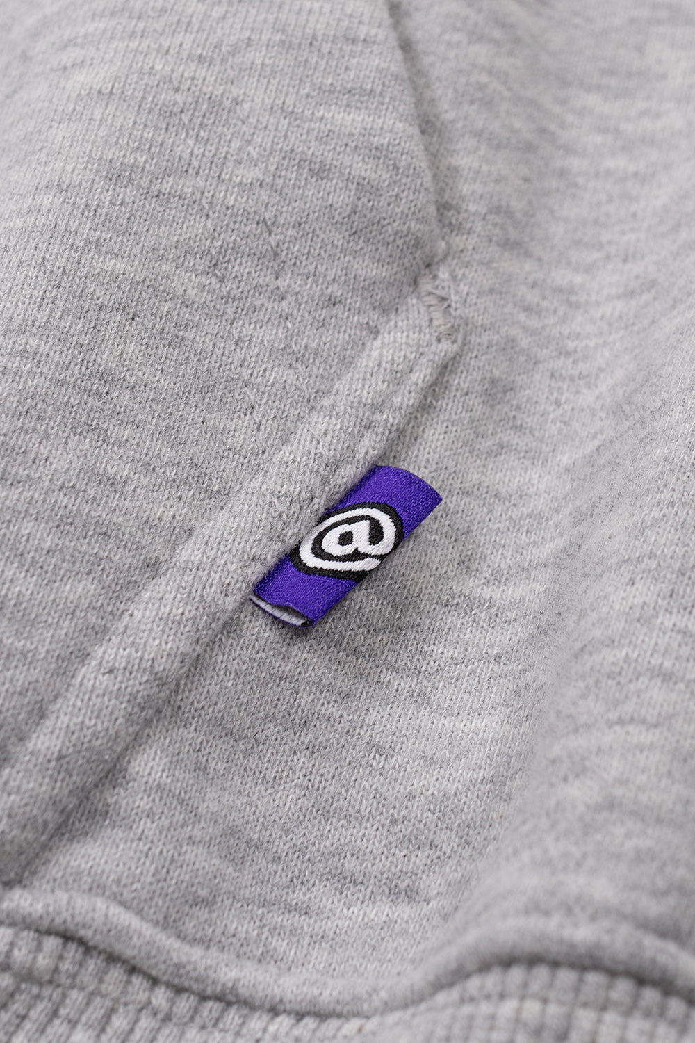 Macro detail of the purple folded woven tag stitched onto the side seam of the kangaroo front pocket, showcasing the signature white embroidered Always "@" spiral logo against the textured heather grey cotton fleece fabric.