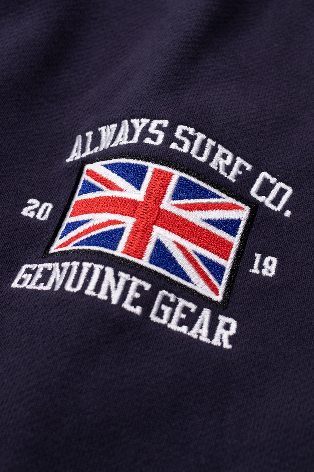 Extreme close-up shot of the left sleeve embroidery on dark navy cotton, depicting the British Union Jack flag framed by white stitched lettering reading "Always Surf Co. 2019 Genuine Gear".