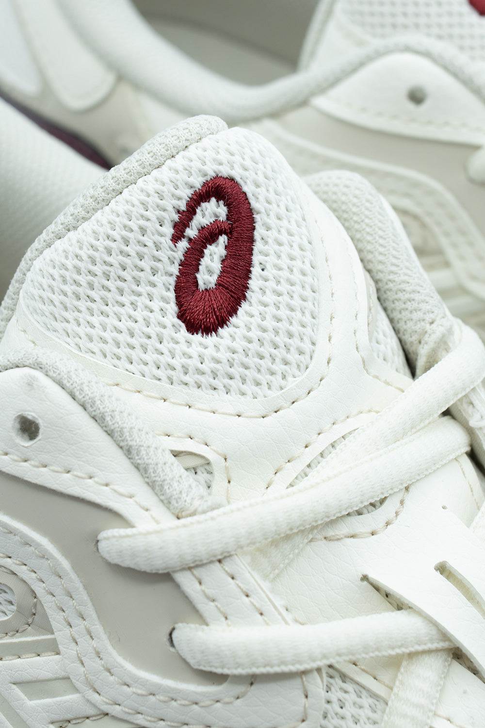 Detailed macro shot of the tongue on the Asics Gel-NYC sneaker, showcasing the textured open-cell cream mesh fabric, neatly stitched collar piping, flat laces, and dimensional dark red embroidered Asics swirl logo