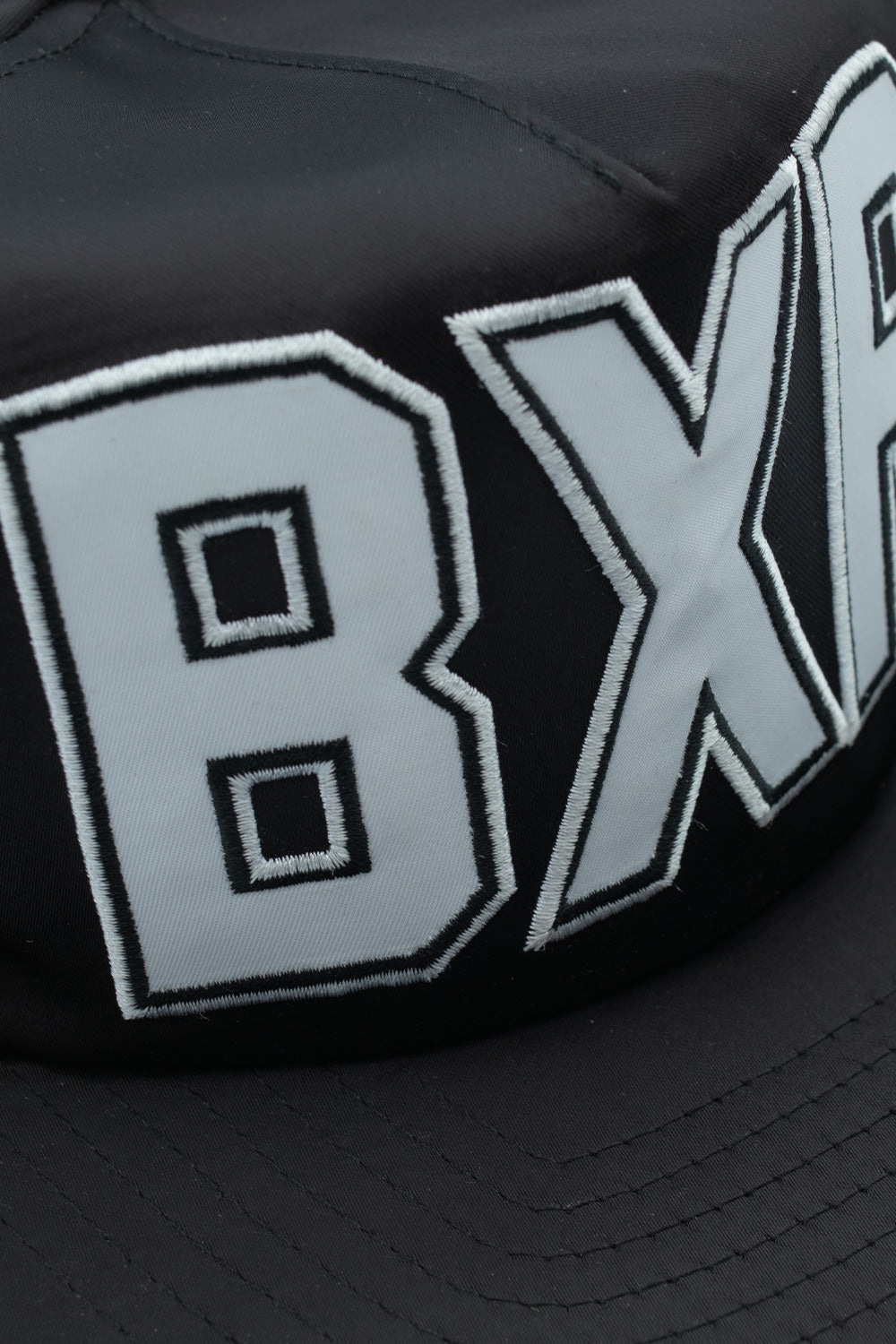 Close-up macro detail of the shiny black polyester upper material and the high-density silver-white embroidered BXR lettering on the cap brim.