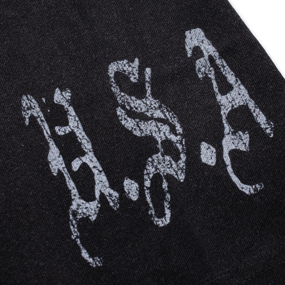 Macro close-up shot of the sleeve cuff graphic on the Born X Raised Llorona T-Shirt, showcasing the distressed, crackle-textured grey screen print of thorny Old English lettering spelling out "U.S.A." on the heavyweight washed black cotton fabric.
