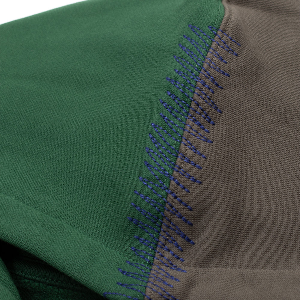 Extreme macro close-up of the top hood seam on the Born X Raised Mashup Zip-Up Hoodie, showcasing the thick decorative purple zigzag whipstitch embroidery joining the split forest green and washed brown fleece fabric panels.