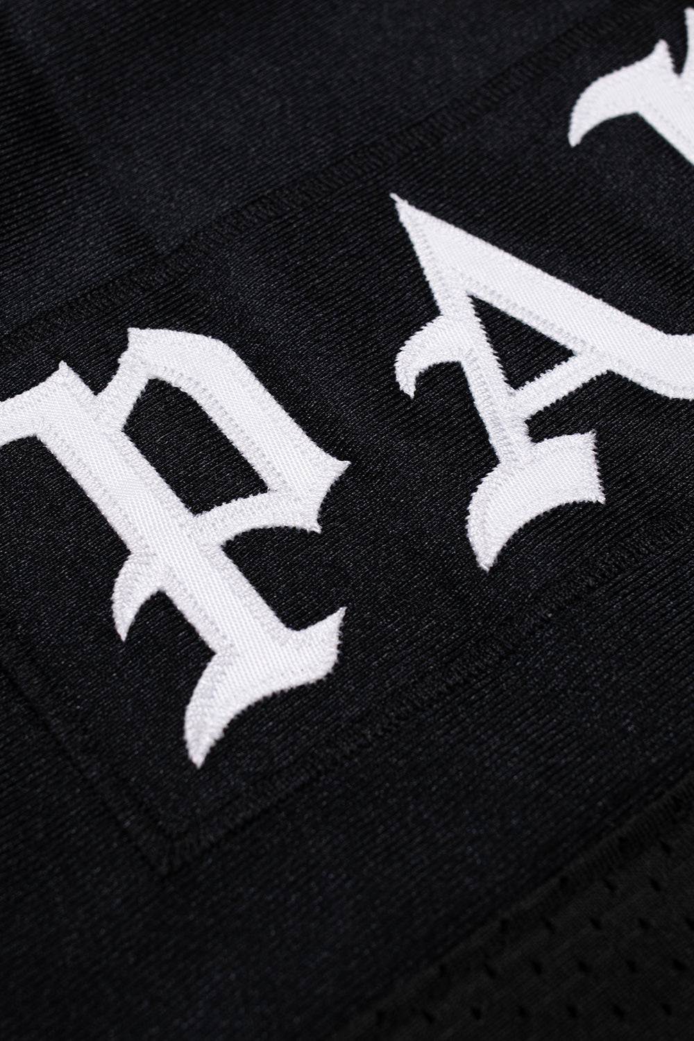 Close-up detail of the white high-density Old English embroidered lettering stitched onto the solid shoulder panel of the black jersey.