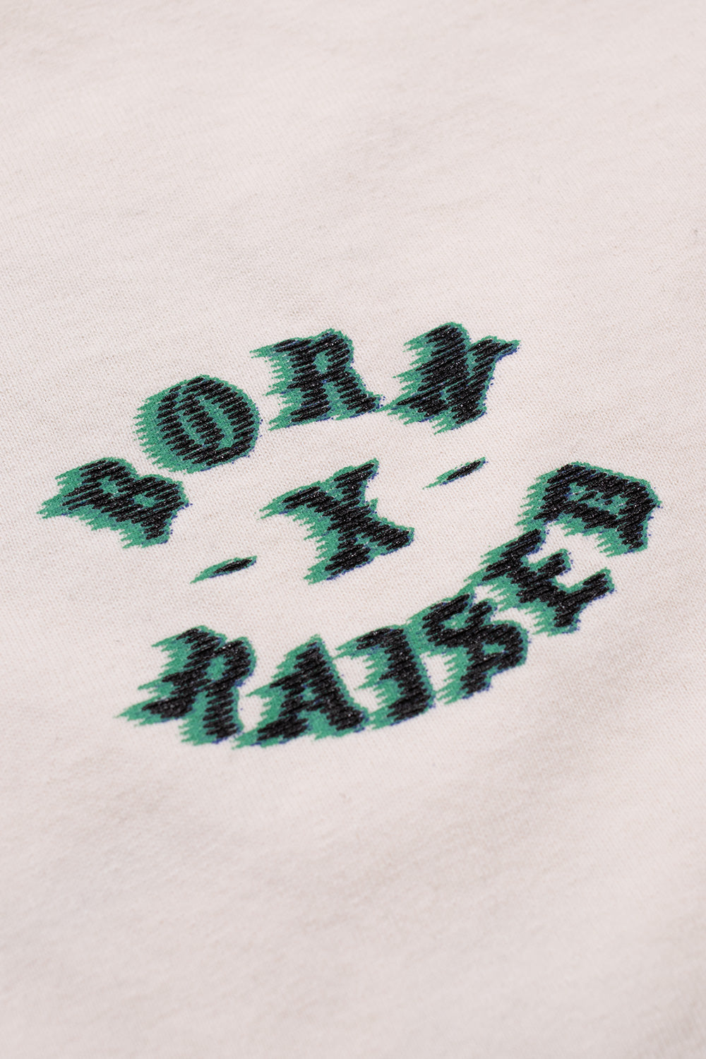 Close-up detail of the green and black speed-blur effect printed Born X Raised logo graphic on the soft cream cotton fabric.