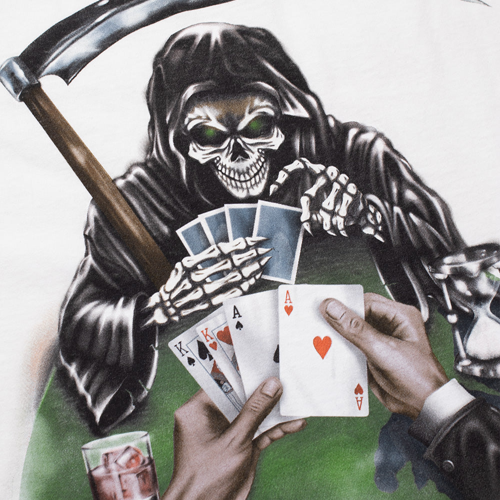 Detailed macro perspective of the back graphic on the Born X Raised Two Pair T-Shirt, capturing the intricate artwork of the skeletal Grim Reaper with glowing green eye sockets holding blue-backed cards, competing against hands holding the Ace of Hearts, Ace of Spades, and King of Spades above a green felt poker table with a drink and hourglass.