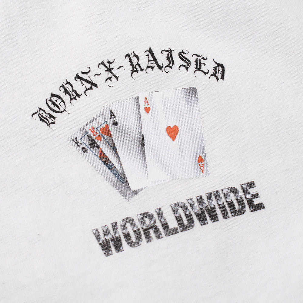 Macro studio close-up of the front chest graphic on the white Born X Raised Two Pair T-Shirt, highlighting the halftone screen-printed poker hand displaying two pair aces and kings, topped by arched black Gothic "BORN-X-RAISED" lettering and underlined by distressed chrome-textured "WORLDWIDE" typography on crisp white cotton jersey.