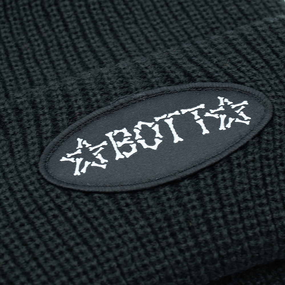 Detailed macro studio close-up of the fold-over cuff on the BoTT Bones Cuff Beanie, capturing the deep elasticated ribbing and the oval black fabric patch stitched with dense white thread forming the skeleton bone "BoTT" logo flanked by bone stars.