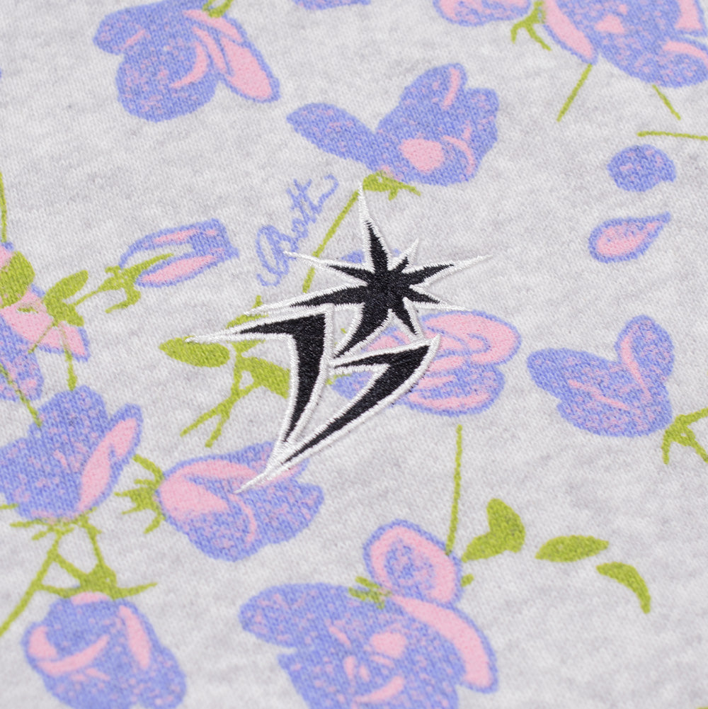 Detailed macro perspective of the chest embroidery on the BoTT Flower Zip Hoodie, capturing the high-density black satin-stitch embroidery with white edge outline forming the signature geometric lightning star "B" emblem, set over the watercolor-style lavender roses and green leaves on ash heather fleece.