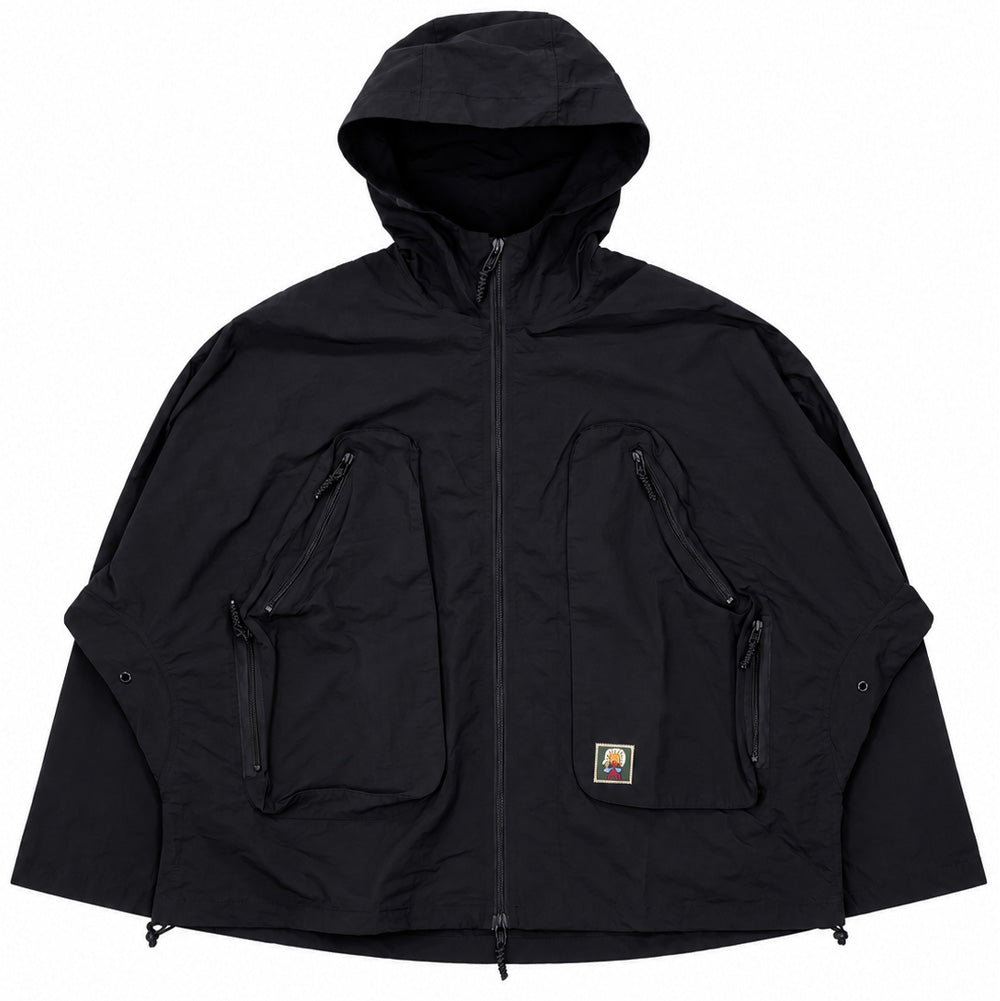 Front flat lay studio view of the Brain Dead Dolman Sleeve Jacket in black, displaying an oversized technical streetwear shell jacket with wide, seamless batwing sleeves, integrated protective hood, full two-way front zipper, two massive multi-entry 3D chest cargo pockets with angled zippers and cord pullers, adjustable snap cuffs, hemline cinch cords, and an embroidered Brain Dead head logo patch on the left pocket.