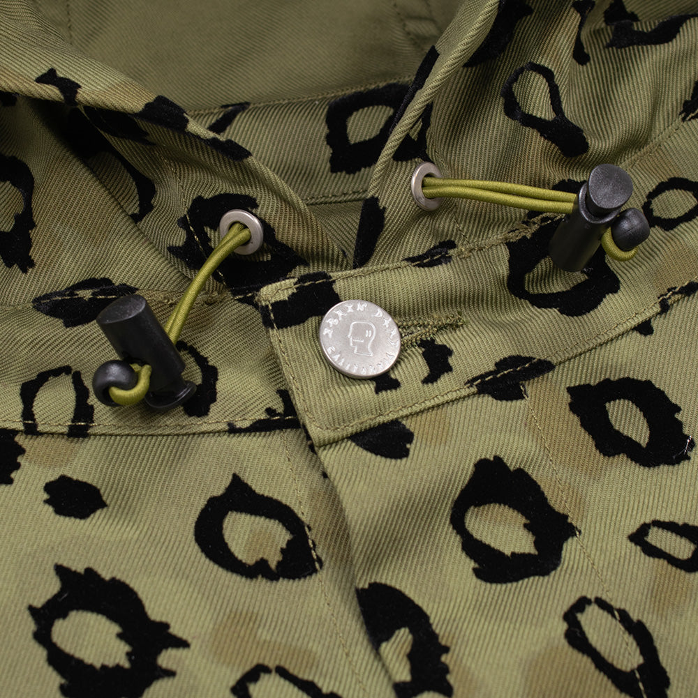 Detailed macro studio close-up of the hood neckline and collar on the Brain Dead Leopard Chore Coat, highlighting the round silver snap button embossed with the Brain Dead head logo and "CALIFORNIA" lettering, beside metallic eyelets, olive bungee drawcords, and black plastic toggle barrel locks against the diagonal twill canvas weave.