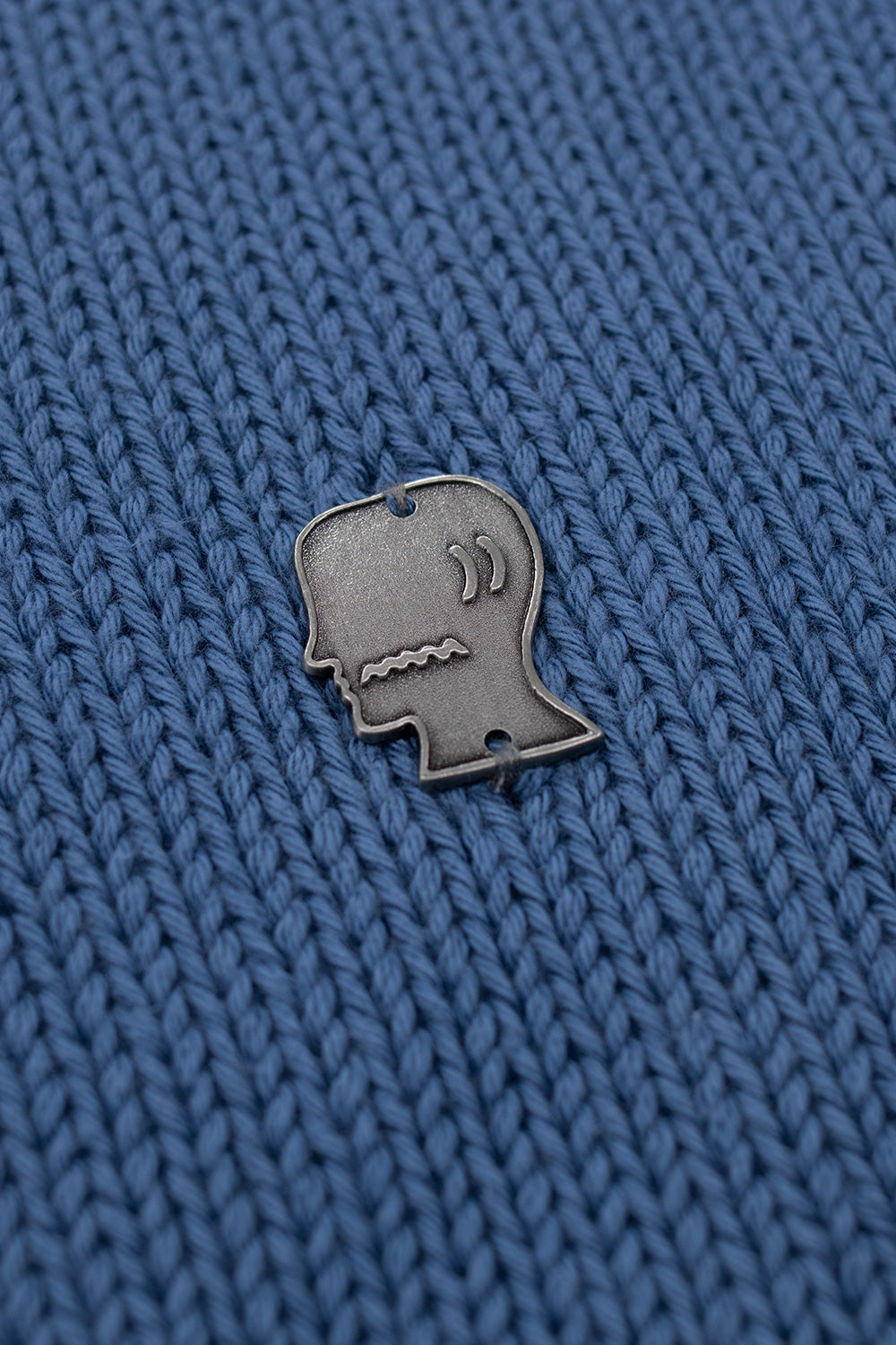 Brain Dead metal logo patch detail on blue turtle neck sweater highlighting signature metal emblem on loosely woven pima cotton streetwear knit made in Peru