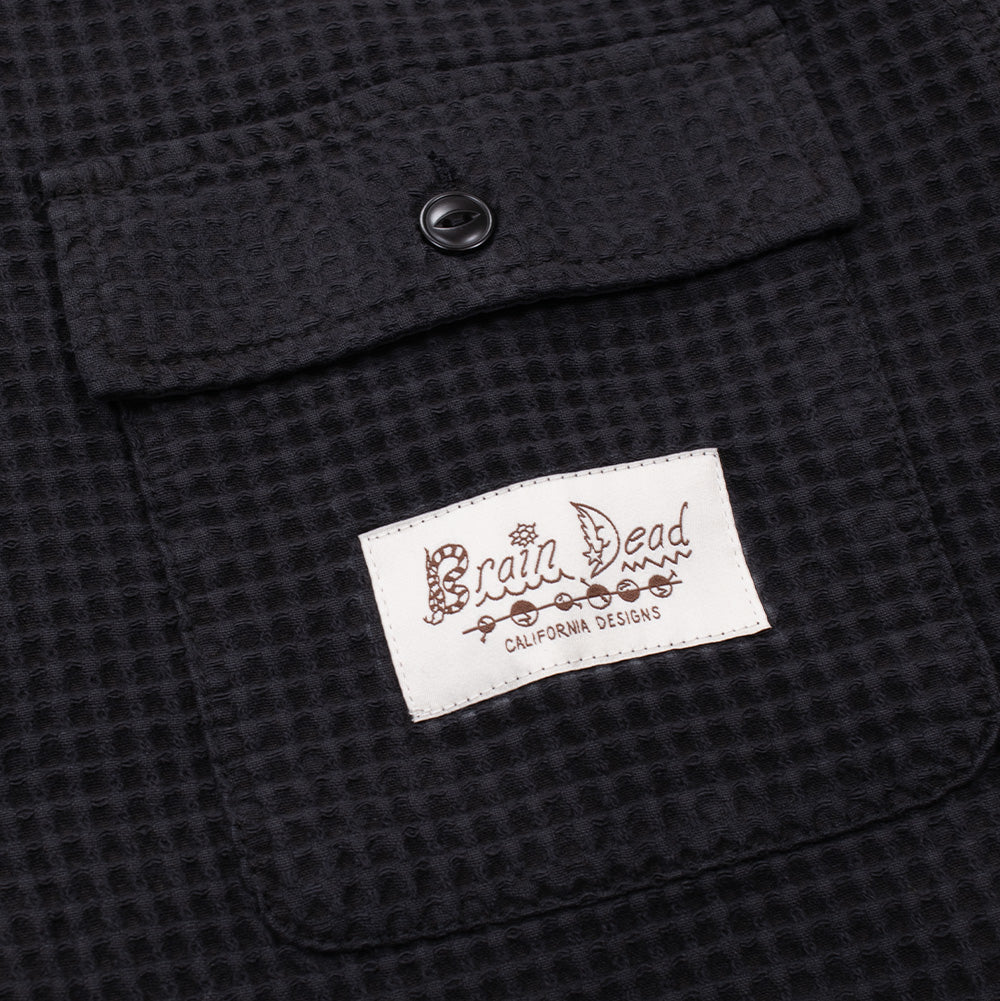 Macro view of the left chest flap pocket on the Brain Dead Waffle Shirt, detailing the single-button flap closure and the stitched cream woven brand patch featuring artistic black illustrated typography reading "Brain Dead" with fish and flower iconography above "CALIFORNIA DESIGNS" lettering.