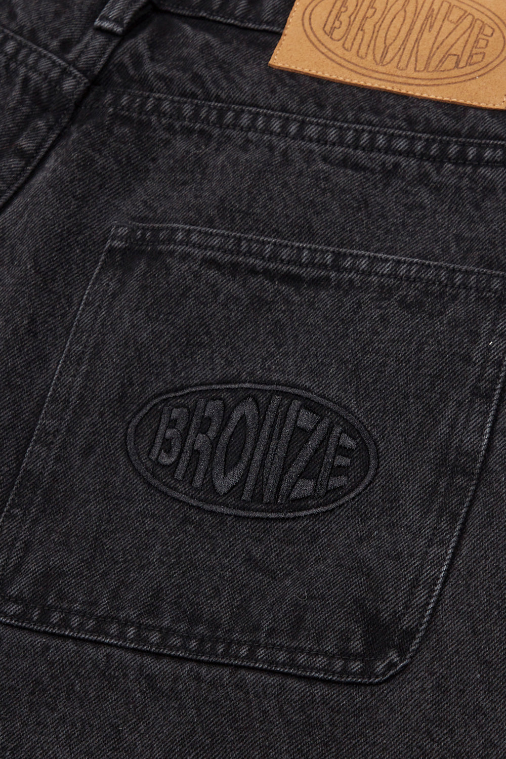 "Detailaufnahme der Bronze 56K Denim Jorts – markantes Logo-Patch auf strapazierfähigem schwarzem Denim"