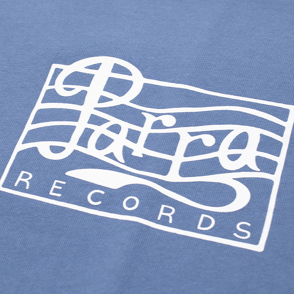 Macro studio view of the front chest graphic on the blue By Parra Records T-Shirt, capturing the clean single-color white ink print featuring the framed cursive "Parra" logo intersecting wavy treble lines above "RECORDS" text on cornflower blue cotton fabric.