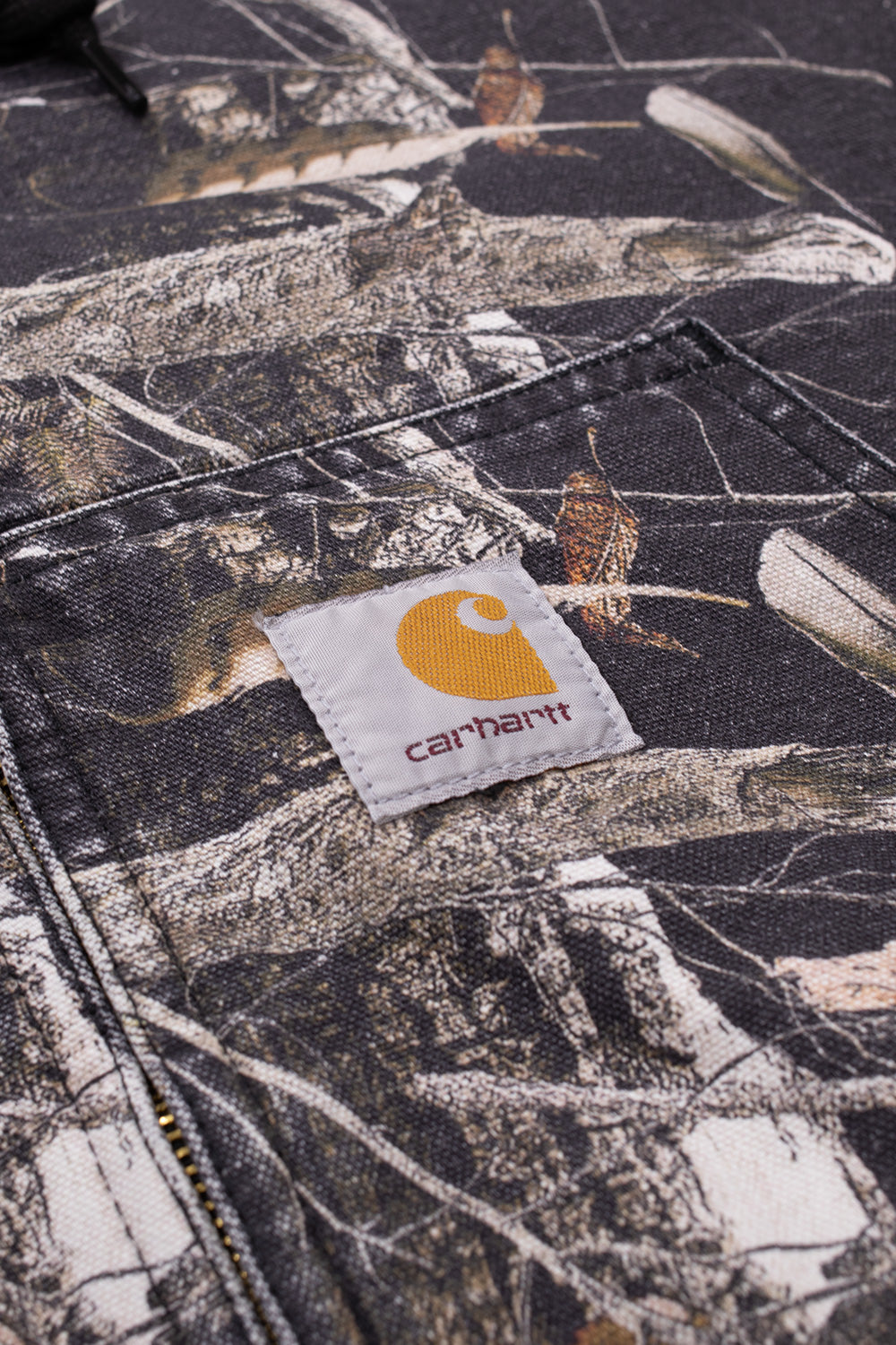 Detailed studio close-up of the front kangaroo pocket on the Carhartt WIP OG Active Jacket, showcasing the iconic white woven square Carhartt label with the golden-yellow "C" emblem and brown brand text, stitched onto the heavy stone-washed Camo Feather Tree cotton canvas with reinforced triple-stitched pocket seams.