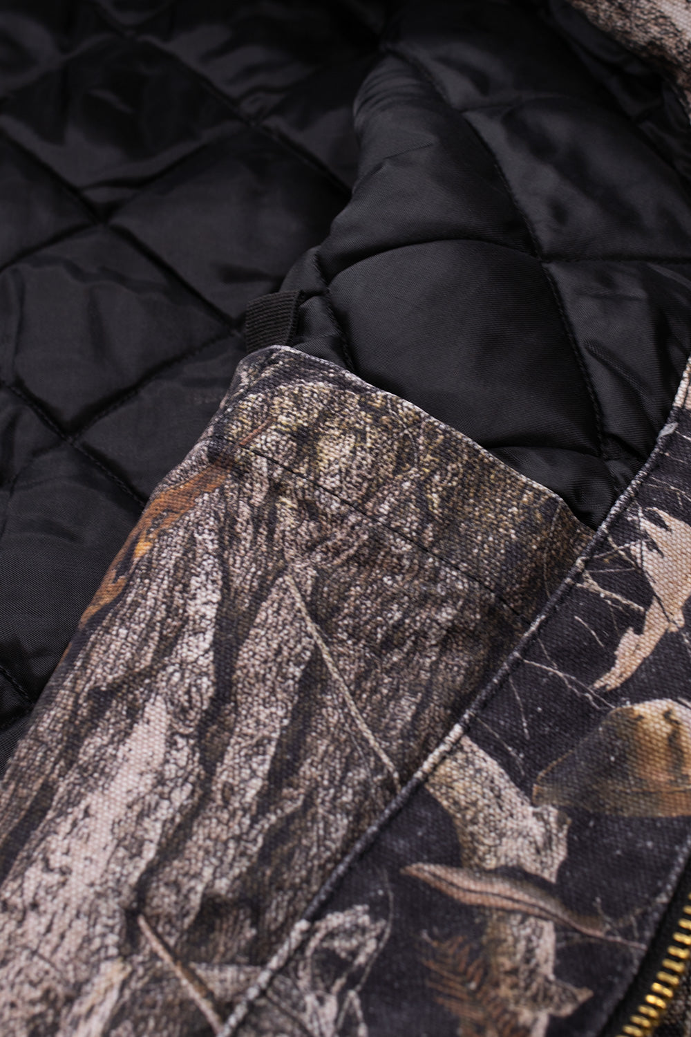 Interior construction macro shot of the Carhartt WIP OG Active Jacket, revealing the durable black diamond-quilted padded lining for warmth, internal patch pocket with bar-tack reinforcement, sturdy brass zipper tape, and the inner facing of the Camo Feather Tree Dearborn canvas shell.