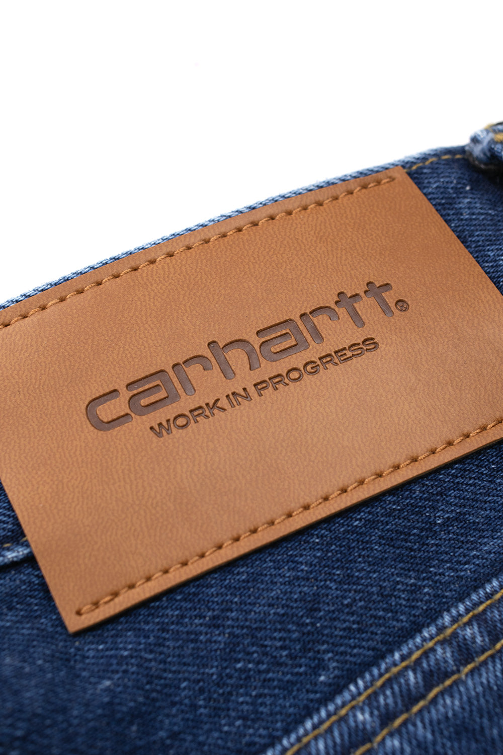 Macro close-up shot of the rear right waistband on the Stone Washed Carhartt WIP Payson Pant, highlighting the textured light brown leather patch debossed with the Carhartt Work In Progress wordmark logo beside double-needle contrast yellow stitching on faded blue denim.