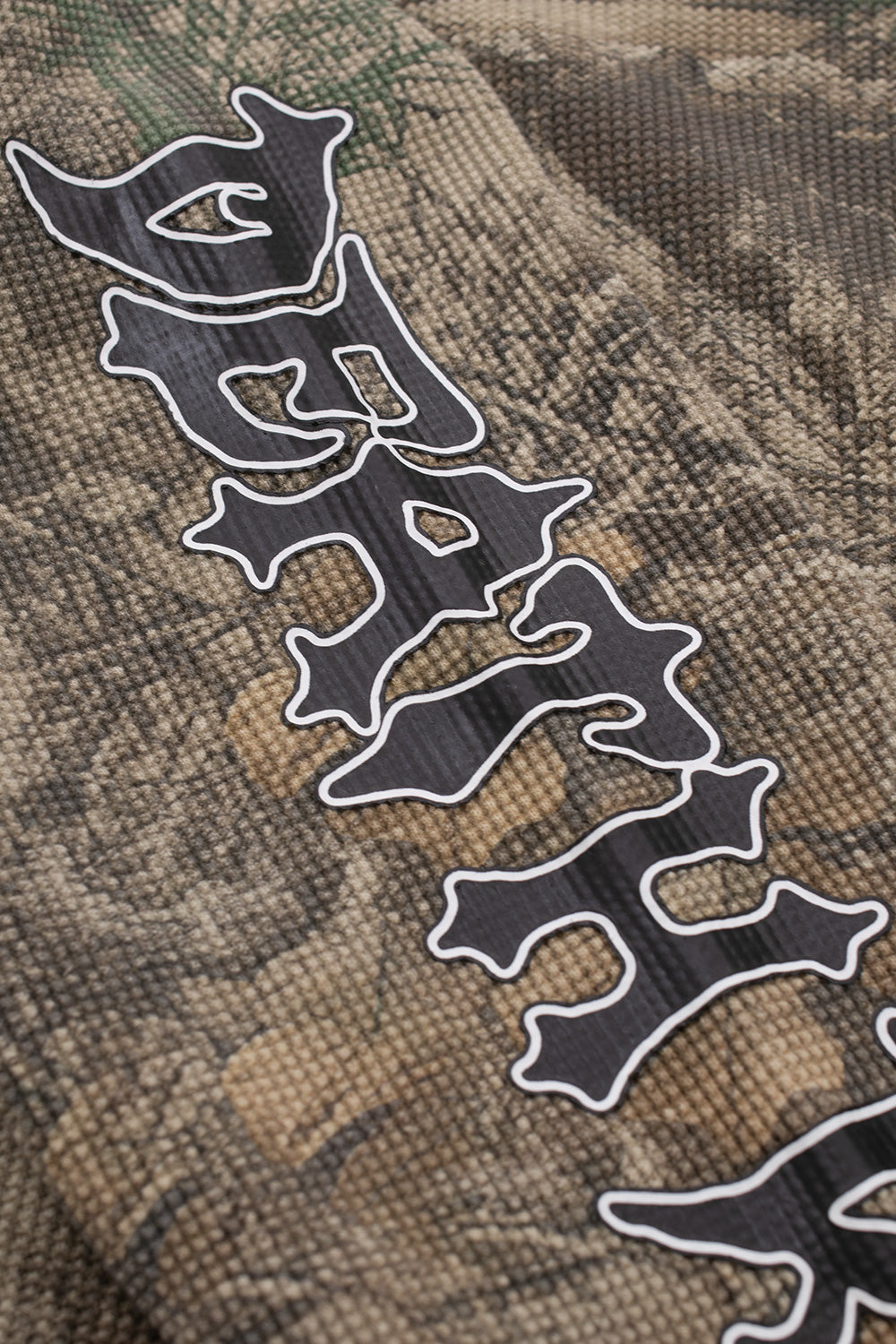 Close-up graphic detail of the stylized black and white metal-style font logo printed over the textured tree camouflage waffle knit fabric.
