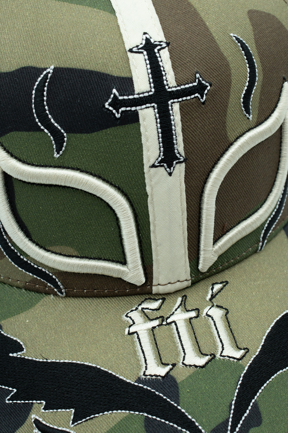 Macro close-up shot of the front embroidery on the Fuck This Industry Mystery Hat, detailing the raised cream satin stitching outlining the eye cutouts, the central embroidered black Gothic cross, and white "fti" Old English typography against the durable cotton camouflage twill fabric