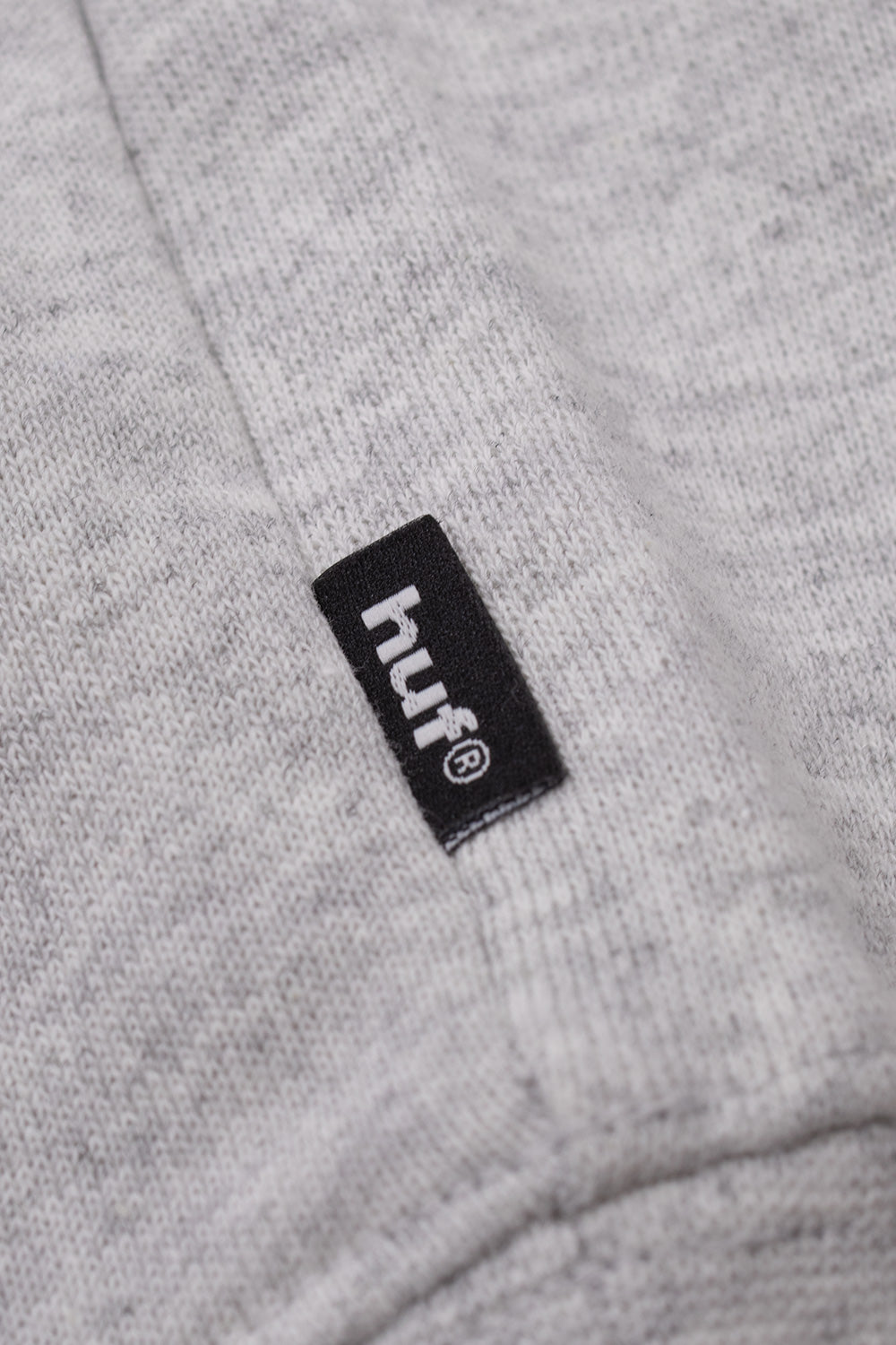 Macro close-up shot of the woven black HUF brand flag label with white registered logo lettering, neatly sewn into the vertical side seam of the heather grey cotton fleece pullover sweatshirt