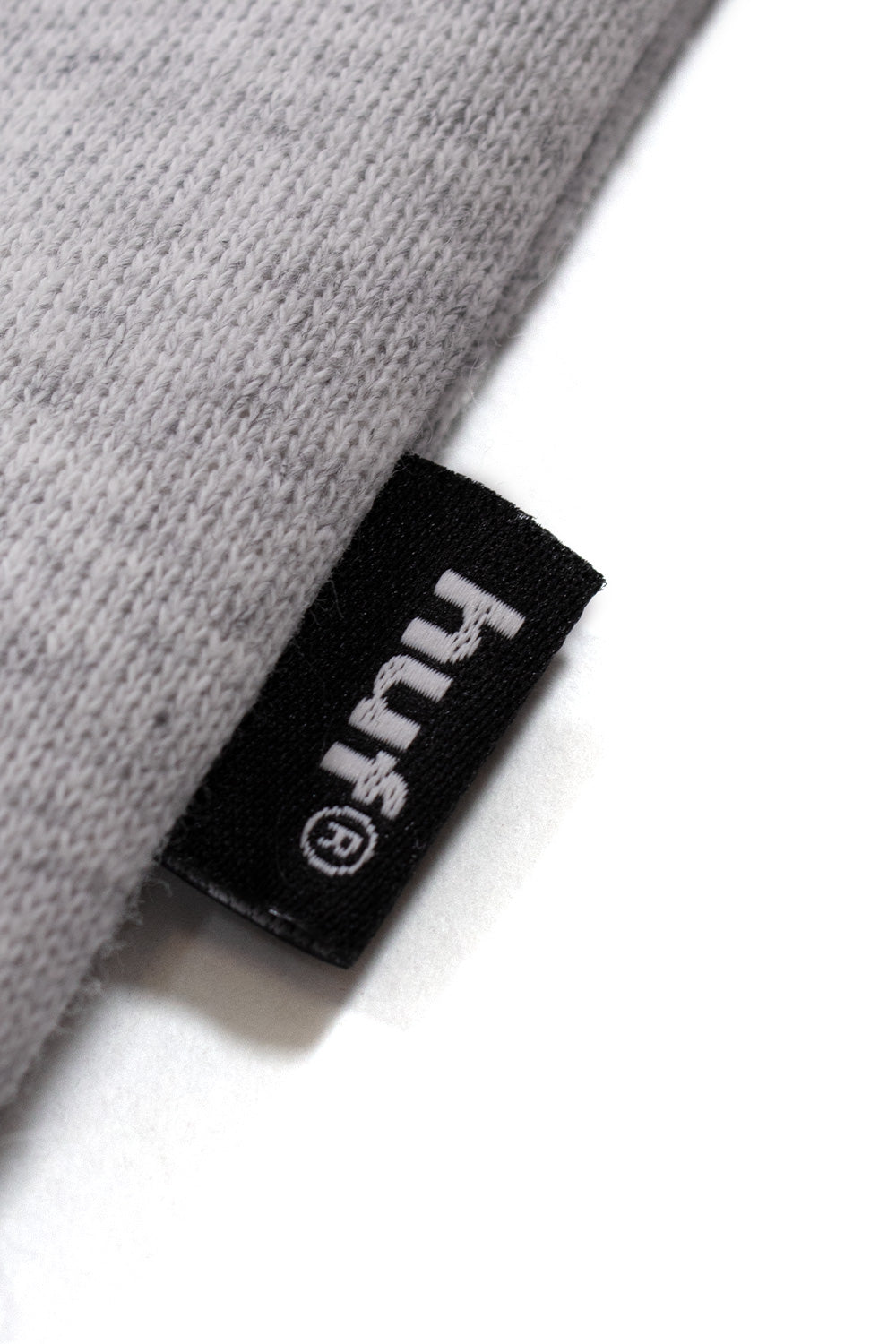 Macro detail shot of the side seam hem on the heather grey cotton-poly fleece sweatshirt, displaying a small black woven tag embroidered with the HUF text logo in white.