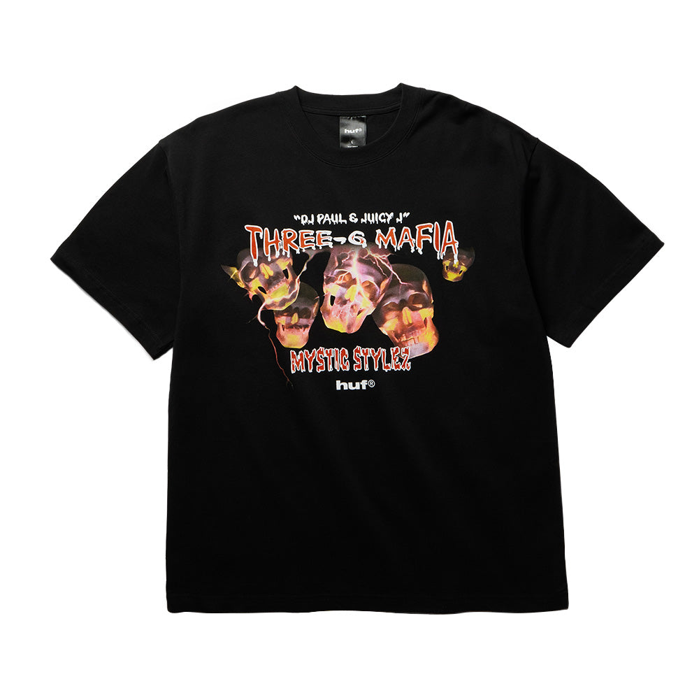 Front flat lay view of the black Huf X Three 6 Mafia Mystic Styles T-Shirt, showcasing a relaxed short-sleeve cut and an intense front graphic print featuring glowing fire-lit skulls engulfed in flames and lightning, framed by white dripping text reading "DJ PAUL & JUICY J" and "THREE-6 MAFIA" above distressed "MYSTIC STYLEZ" lettering and the classic "huf®" logo.