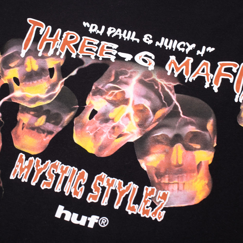 Macro studio close-up of the front chest graphic on the Huf X Three 6 Mafia Mystic Styles T-Shirt, highlighting the detailed fiery orange and yellow skulls, jagged electric bolts, dripping white graffiti font for Three-6 Mafia, textured "MYSTIC STYLEZ" typography, and the crisp white HUF brand wordmark on black cotton fabric.