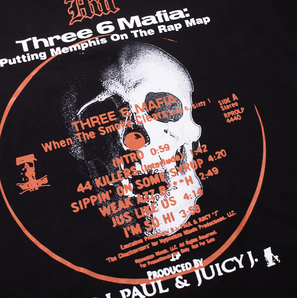 Studio macro perspective of the back print on the Huf X Three 6 Mafia Mystic Styles T-Shirt, detailing the circular orange vinyl label border, album song titles like "Sippin' On Some Syrup" and "Weak Azz Bitch", the printed skull centerpiece, and bold "PRODUCED BY D.J. PAUL & JUICY J." typography on the black shirt.