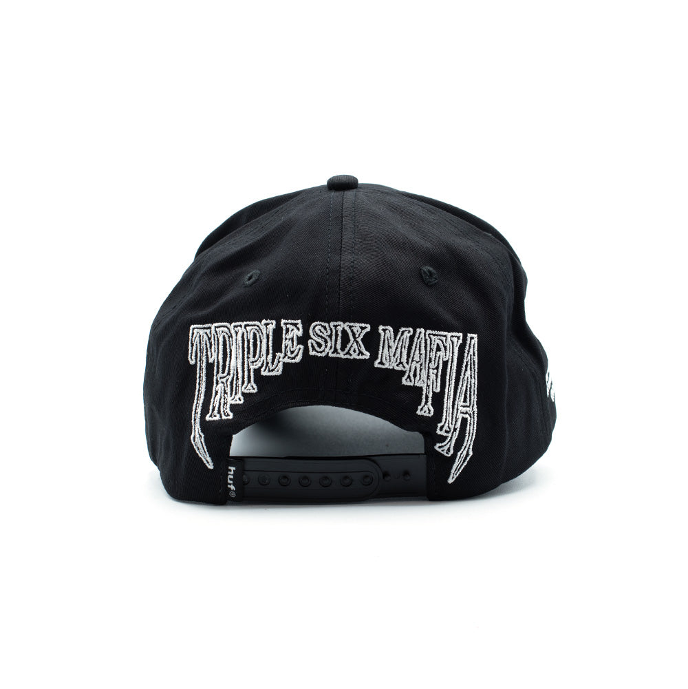 Rear studio perspective of the black HUF x Three 6 Mafia Snapback cap, highlighting the large dripping horror-font white outline embroidery reading "TRIPLE SIX MAFIA" arched over the open rear cutout, complemented by a black adjustable plastic snapback strap and a small woven HUF flag tag.