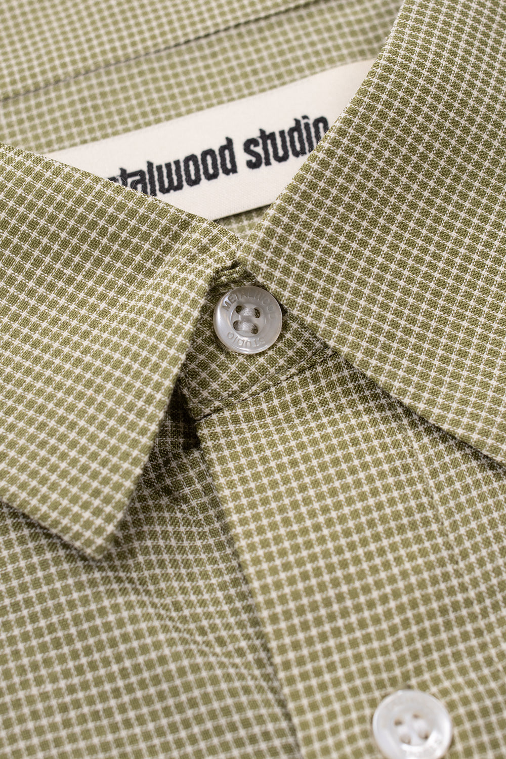 Macro view of the green micro-pattern fabric and Metalwood logo button on an oversized button-up shirt inspired by vintage golf fashion
