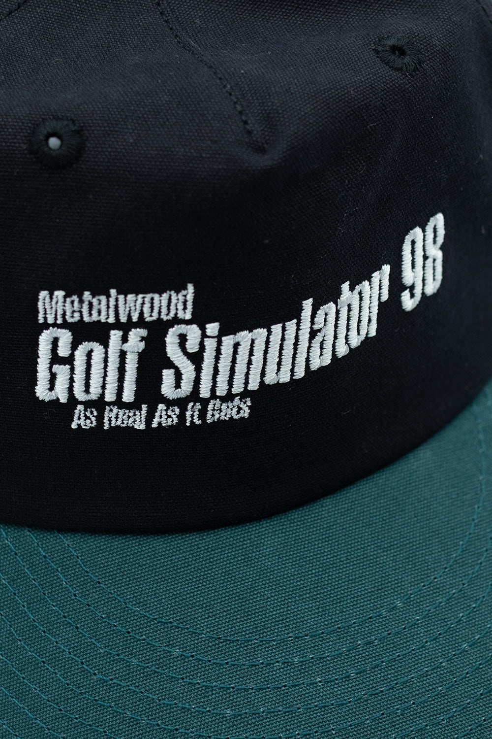 Close-up of the Metalwood Studio Simulator 98 5-panel snapback cap's embroidered logo in vibrant teal and black.