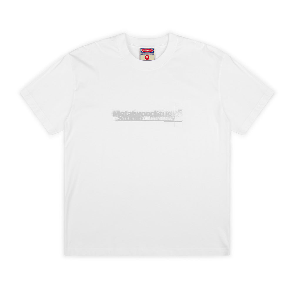 Front flat lay studio view of the Metalwood Studio Skidmark T-Shirt in solid white, showcasing a relaxed short-sleeve streetwear fit crafted from cotton jersey, ribbed crewneck collar with red and blue woven inner brand tag, and a centered chest screen print featuring a distressed, spray-blurred "Metalwood Studio" graphic in light silver-grey.