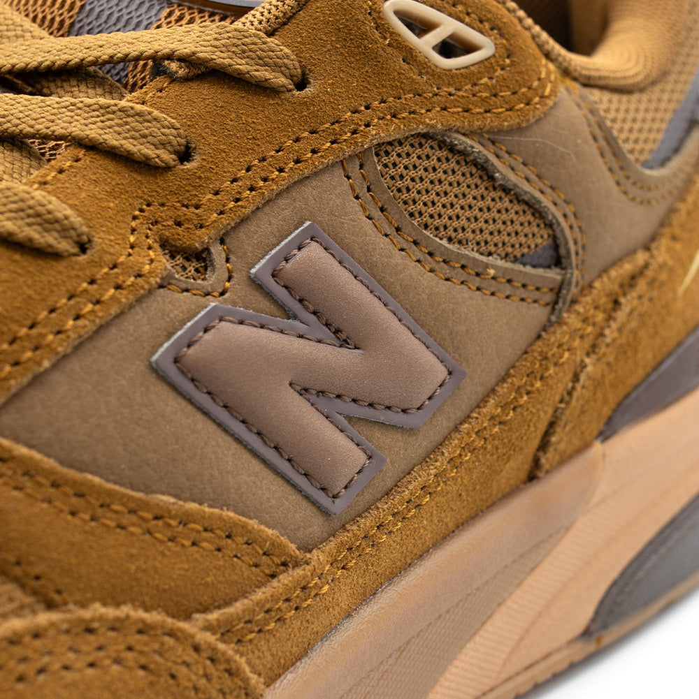 Detailed macro perspective of the lateral quarter panel on the New Balance Numeric Andrew Reynolds 933, showing the smooth tan leather base, double-stitched golden suede eyestay, upper TPU eyelet, and the inset grey leather N logo framed by dark brown contrast border stitching.