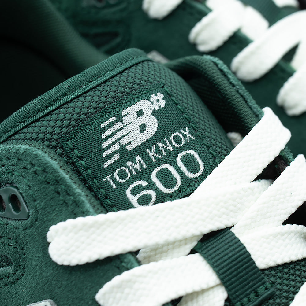 Macro studio close-up of the tongue label on the New Balance Numeric Tom Knox 600, detailing the breathable dark green honeycomb mesh fabric, white cotton laces, and embroidered white NB# Tom Knox 600 logo text.