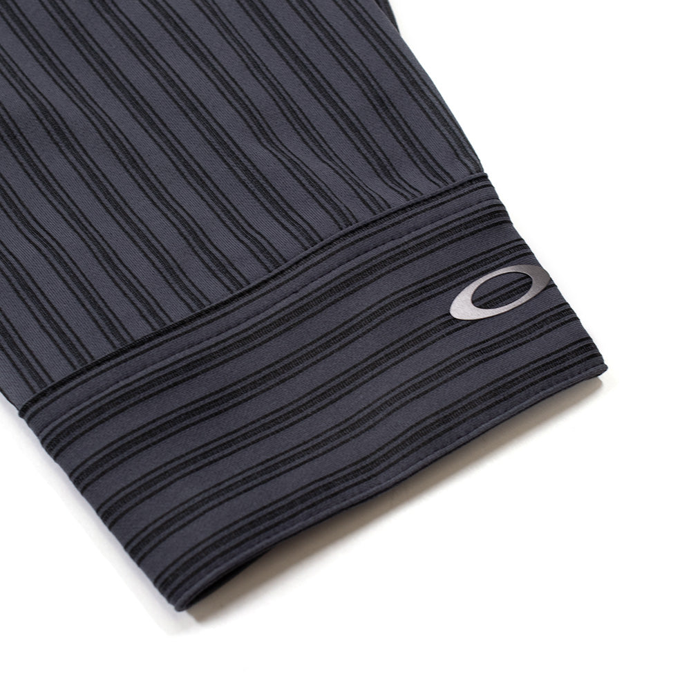 Macro studio close-up of the sleeve cuff on the Oakley FGL Fuel LS Shirt 6.7 in Phantom, detailing the horizontally striped barrel cuff trim, neat edge stitching, and the bonded metallic silver Oakley Ellipse emblem positioned near the wrist edge.
