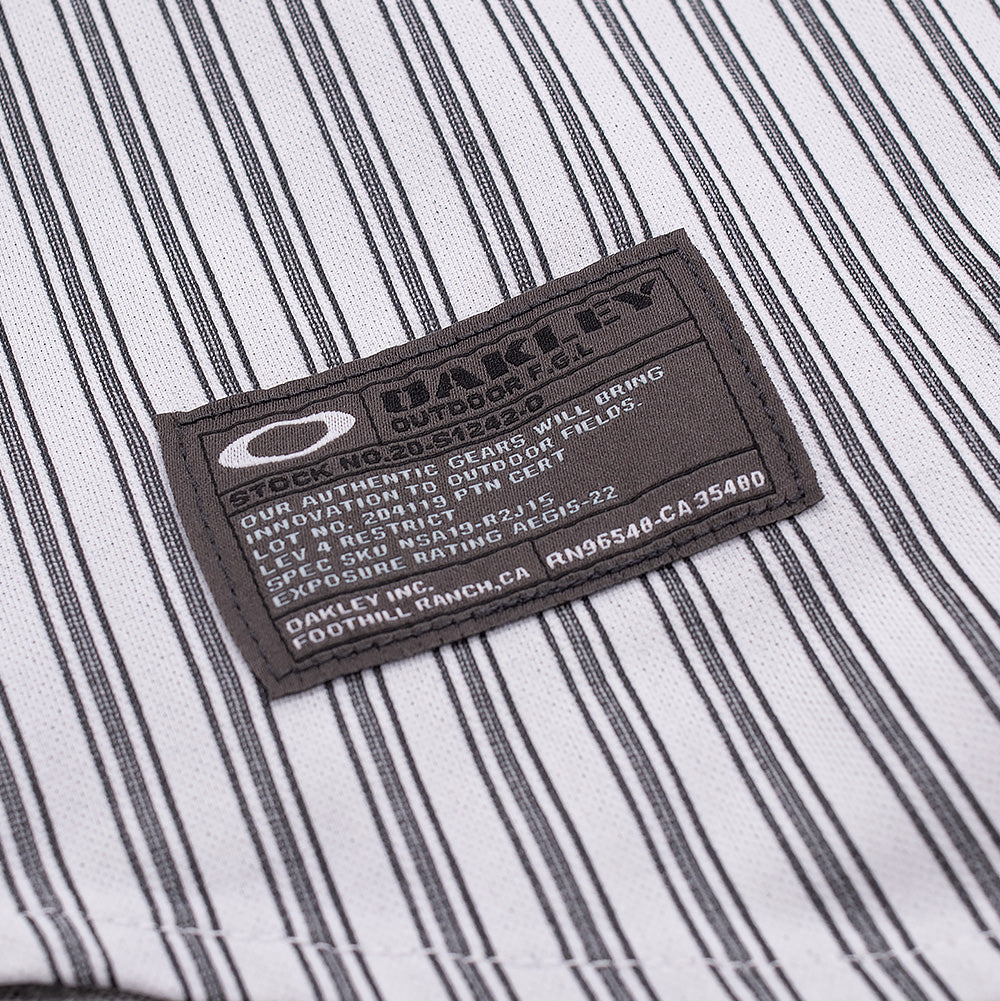 Macro studio view of the lower hem branding on the Oakley FGL Fuel LS Shirt White, focusing on the charcoal woven utility spec tag stitched cleanly onto the striped shell, featuring the white Oakley Ellipse logo and technical specification text.
