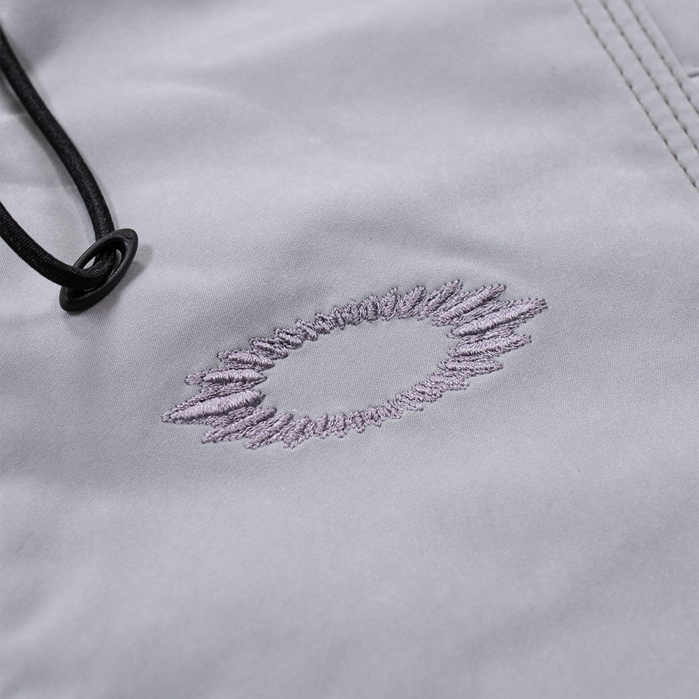 Detailed macro perspective of the chest embroidery on the Oakley FGL Sector Jacket in Winter Grey, capturing the tonal grey satin-stitch embroidery forming the jagged, soundwave-style scratch outline of the Oakley Ellipse emblem set beside curved seamlines and black elastic drawcords.
