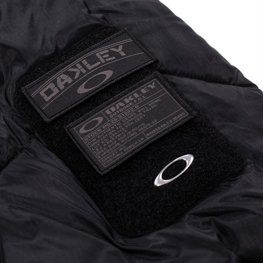 Macro studio view of the modular arm badge system on the Oakley FGL Stealth Ins Jacket 2.0, displaying stacked rectangular velcro hook-and-loop patches with white-and-grey military-spec text, "OAKLEY OUTDOOR F.G.L." insignia, and a metallic silver Oakley Ellipse badge.