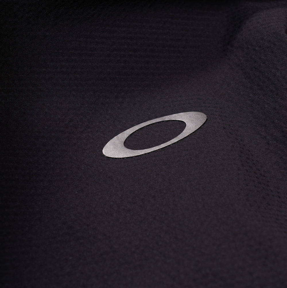 Extreme macro close-up of the rear shoulder branding on the Oakley FGL Stroke Anorak, showcasing the reflective silver metallic heat-transfer Oakley Ellipse emblem cleanly bonded onto the fine-textured, weather-resistant Phantom black technical woven fabric.