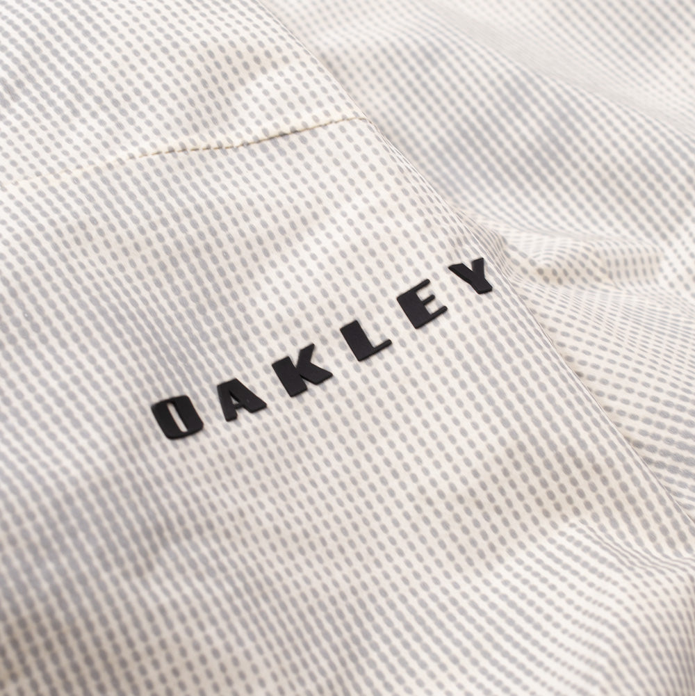 Macro shot of the lower rear hem branding on the Oakley Mthod Glow Shell Jacket, displaying the crisp black uppercase "OAKLEY" text printed across the durable, weather-resistant micro-grid textile.