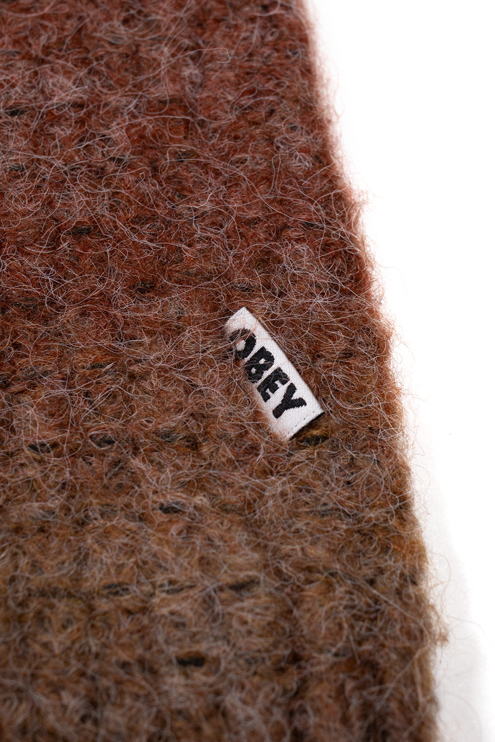Extreme close-up shot of the lower side seam on the brown Obey Anton Cardigan, featuring the small white folded woven tag with black embroidered Obey typography attached to the soft brushed ombré knit fabric.