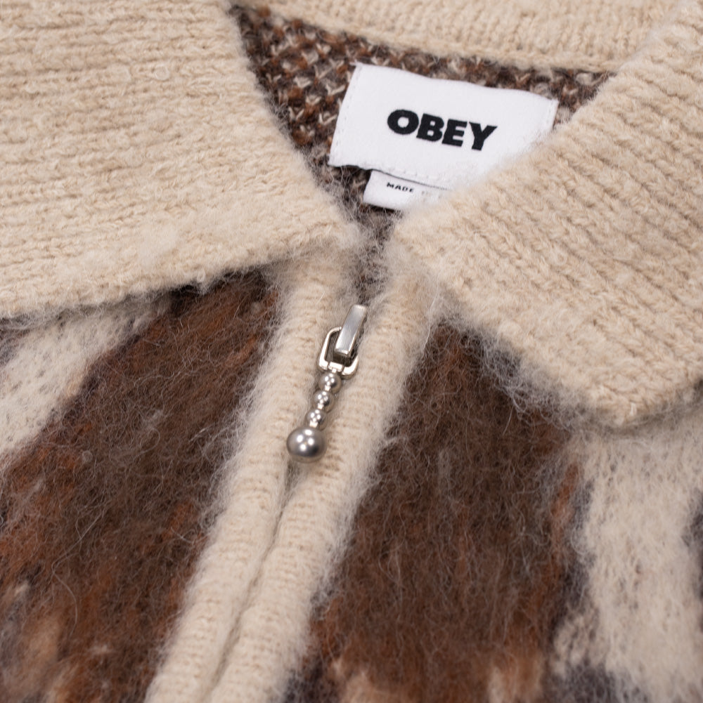 Macro studio close-up of the neckline and zipper on the Obey Cow Pattern Zip Sweater, detailing the ribbed cream knit fold-down collar, stitched white OBEY neck label, and a distinct silver-tone metallic zipper pull shaped like a textured ball chain.