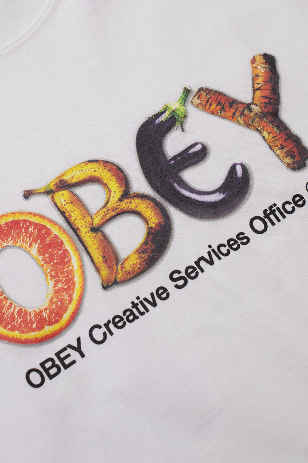 Detailed angled close-up of the front chest graphic on the white Obey tee, highlighting the photographic print of grocery food typography shaped like the Obey wordmark along with the black OBEY Creative Services Office 1989 lettering on white cotton knit fabric.